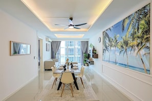 A modern dining area with a ceiling fan, a large window with curtains, and a painting of palm trees. - The Shore Residence Melaka By I Housing (Malacca City)