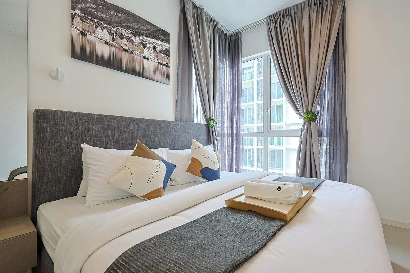 Ein ordentlich bezogenes Bett mit Kissen und einer Ablage für Handtücher, ein großes Fenster mit Vorhängen und ein Bild an der Wand.