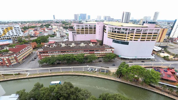 A cityscape with a prominent pink building with a unique curved design, surrounded by other buildings and a river with a bridge. - The Shore Residence Melaka By I Housing (Malacca City)