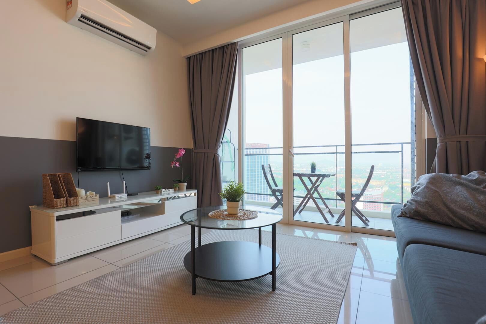 A modern living room with a flat-screen TV, a sofa, a coffee table, and a balcony with a view.