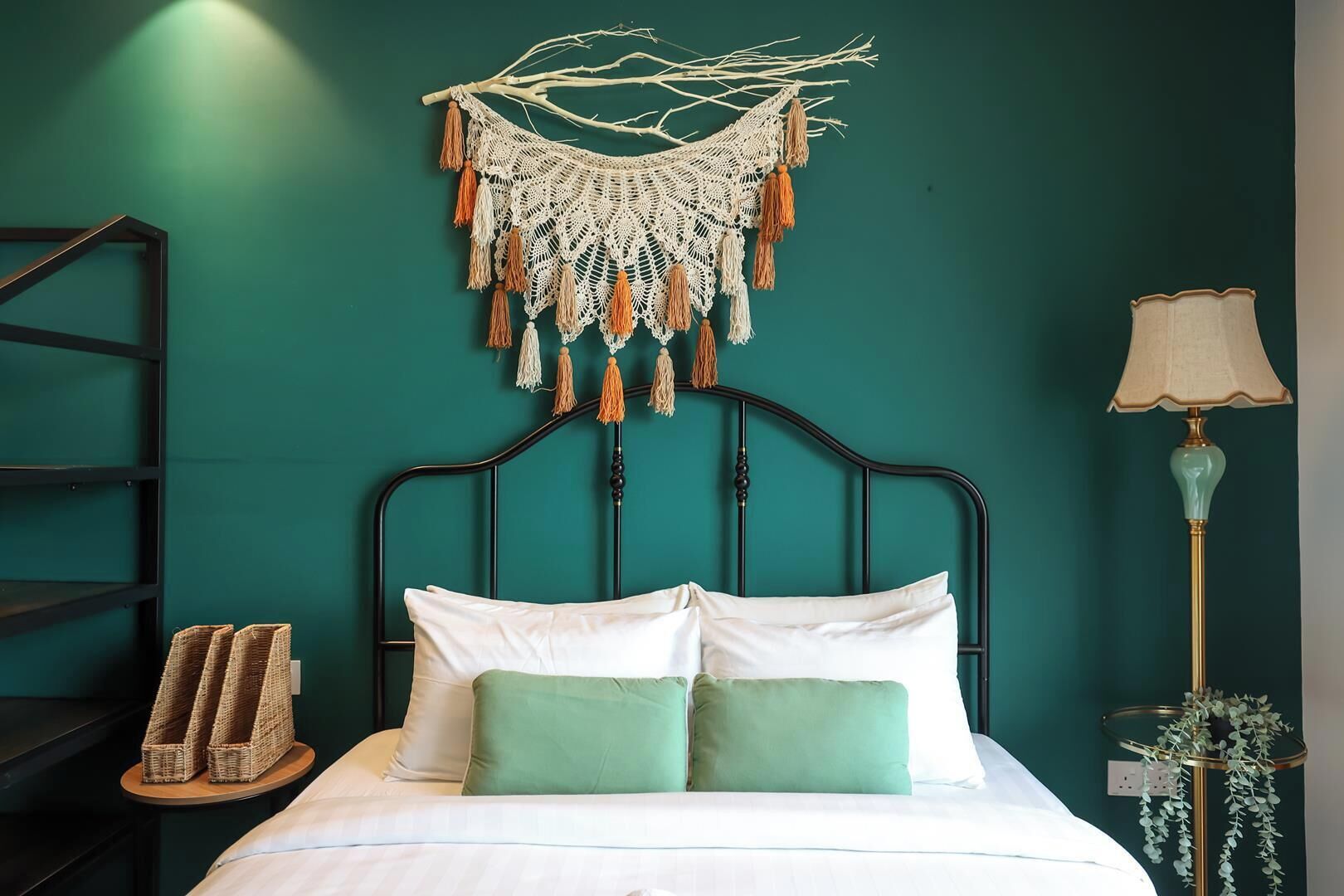 A bed with white and green pillows, a macrame wall hanging, a lamp, and a shelf with woven baskets.