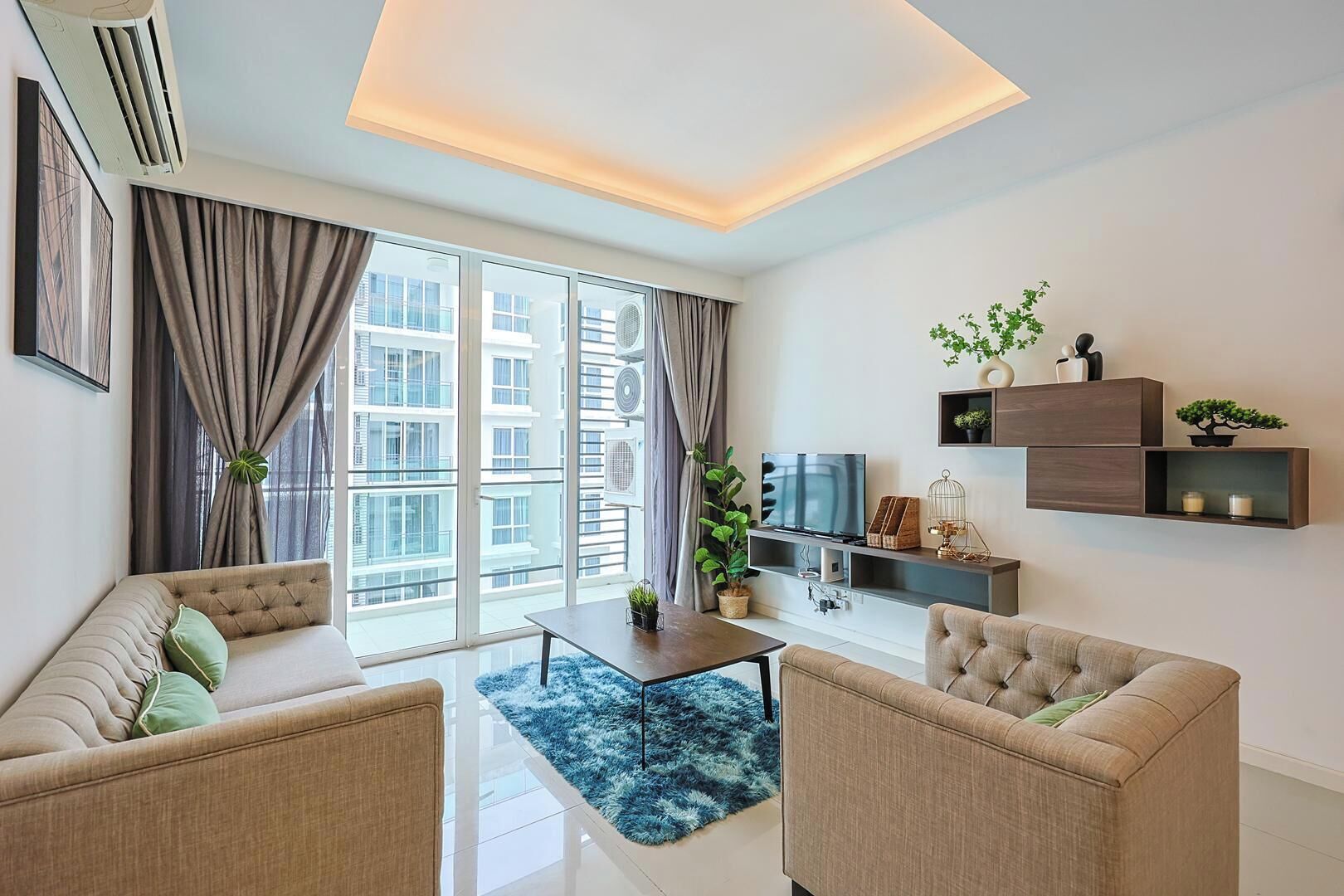 Ein modernes Wohnzimmer mit einer Couch, Sesseln, einem Couchtisch und einem Fernseher. Große Fenster mit Vorhängen, eine Deckenleuchte und eine kleine Pflanze auf einem Regal.