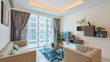 Ein modernes Wohnzimmer mit einer Couch, Sesseln, einem Couchtisch und einem Fernseher. Große Fenster mit Vorhängen, eine Deckenleuchte und eine kleine Pflanze auf einem Regal.