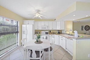 A modern kitchen with white cabinets, a dining table with chairs, a ceiling fan, and a window with blinds.