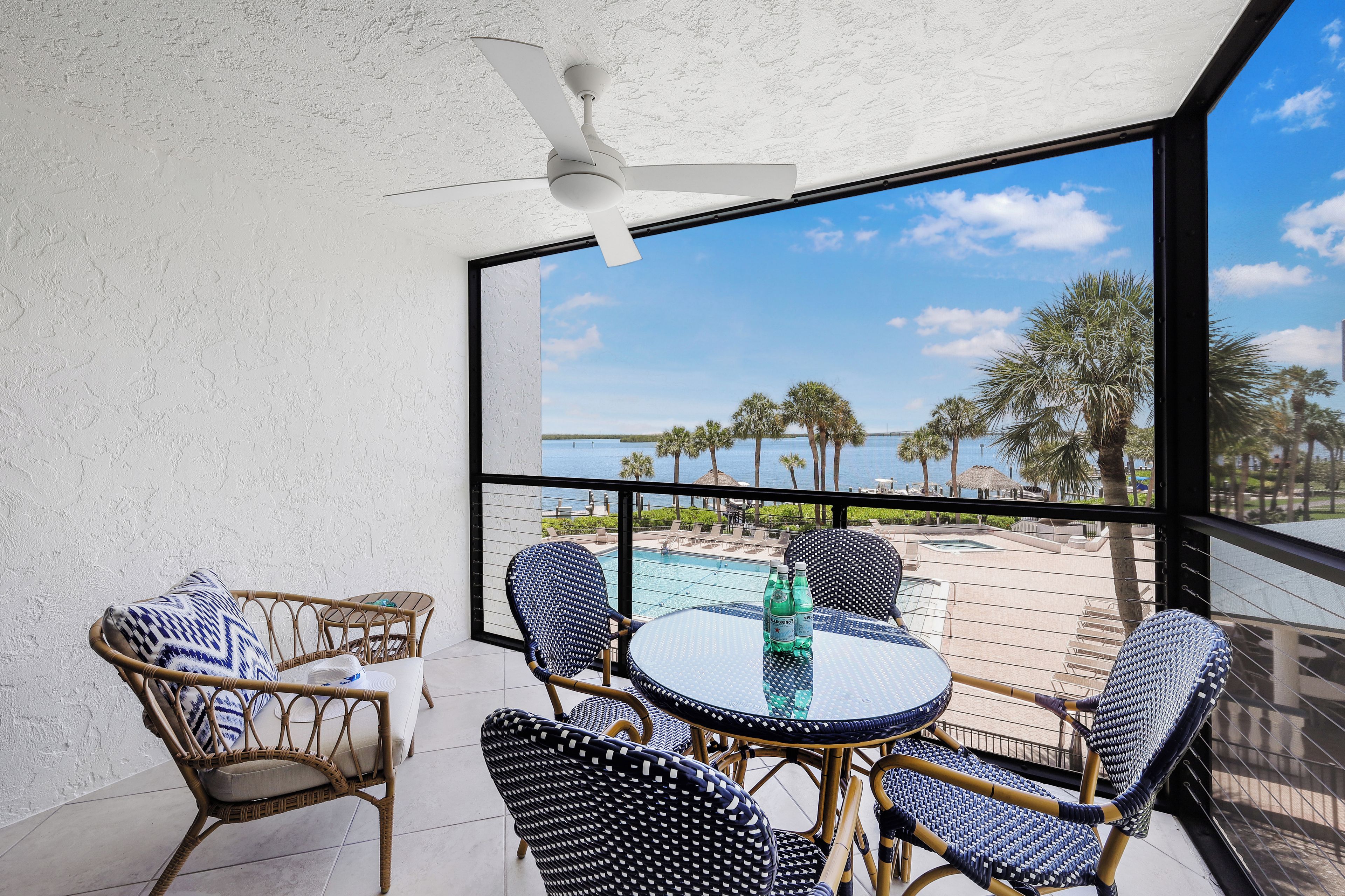 A balcony with wicker furniture, a glass table, and a view of a pool and palm trees.
