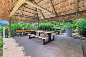 Uma área de estar externa com telhado de palha, piso de concreto e vegetação ao redor.