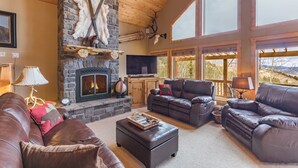 A cozy living room with a stone fireplace, leather sofas, and large windows offering a view of the outdoors.