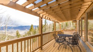 A wooden deck with a pergola, a round table with chairs, and a view of mountains and trees.