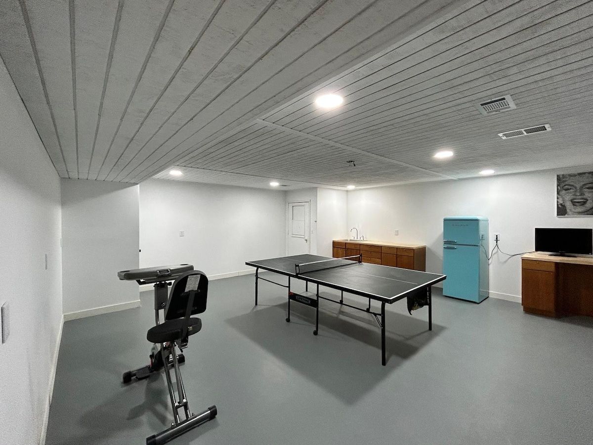 A room with a ping pong table, a small exercise bike, a blue refrigerator, and a wooden desk with a television.