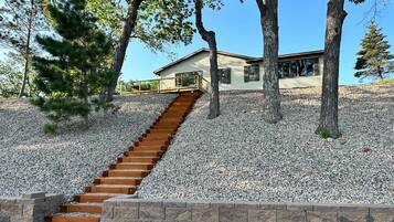 A house with a wooden staircase leading to a deck, surrounded by trees and gravel.