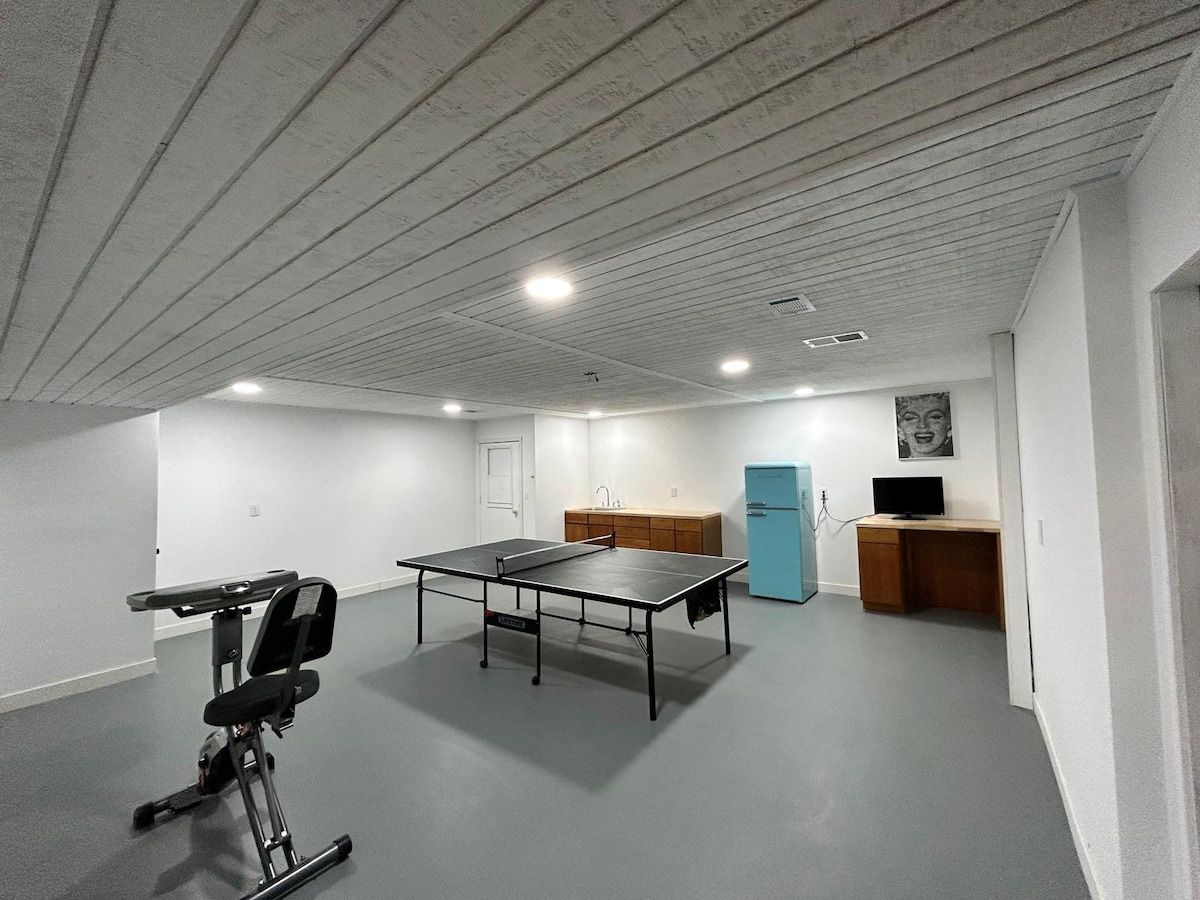 A room with a ping pong table, a small exercise bike, a blue refrigerator, a wooden desk, and a wall-mounted TV.