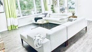 Een moderne woonkamer met een hoekbank, salontafel en vloerlamp. Er zijn grote ramen met gordijnen, een plafondventilator en uitzicht op buiten.