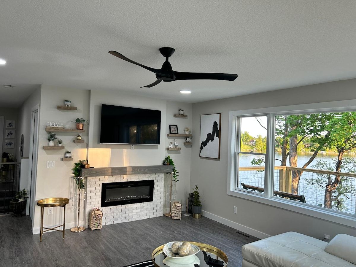 A living room with a fireplace, a ceiling fan, a TV mounted on the wall, and a window with a view of a body of water.