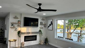 A living room with a fireplace, a ceiling fan, a TV mounted on the wall, and a window with a view of a body of water.