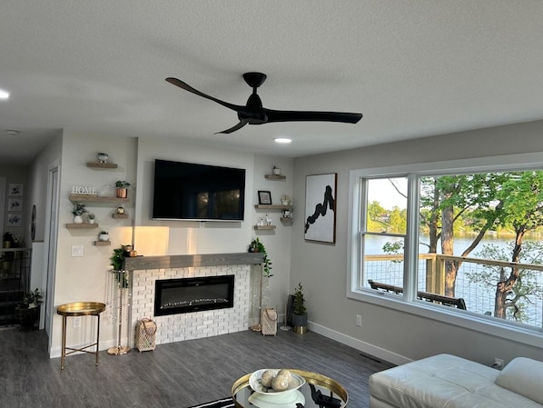 Una sala de estar con chimenea, ventilador de techo, televisor montado en la pared y ventana con vista a un cuerpo de agua.