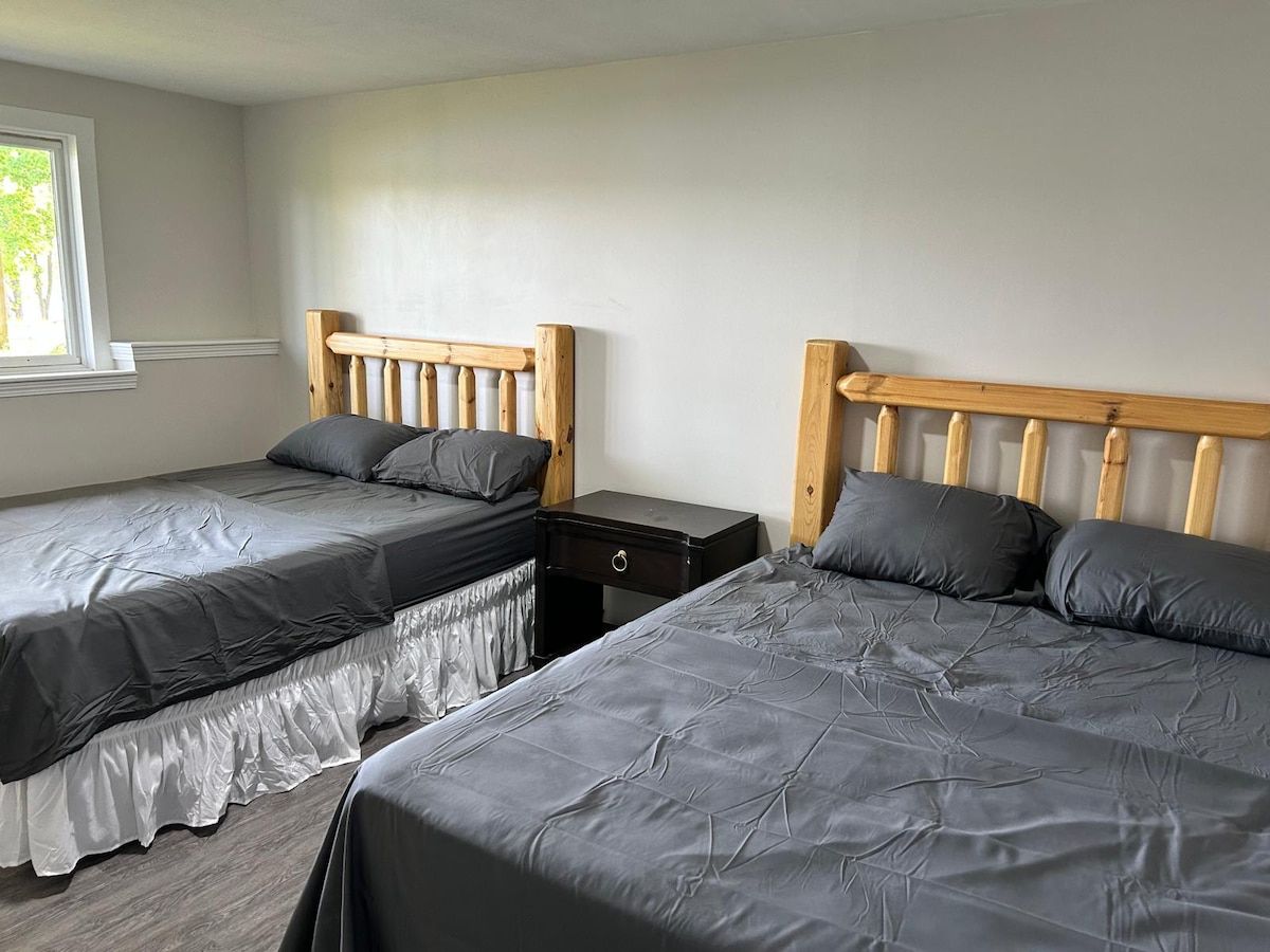 Une chambre avec deux lits, chacun doté d’une tête de lit en bois et d’une literie grise. Une petite table de chevet avec un tiroir se trouve entre les deux lits.
