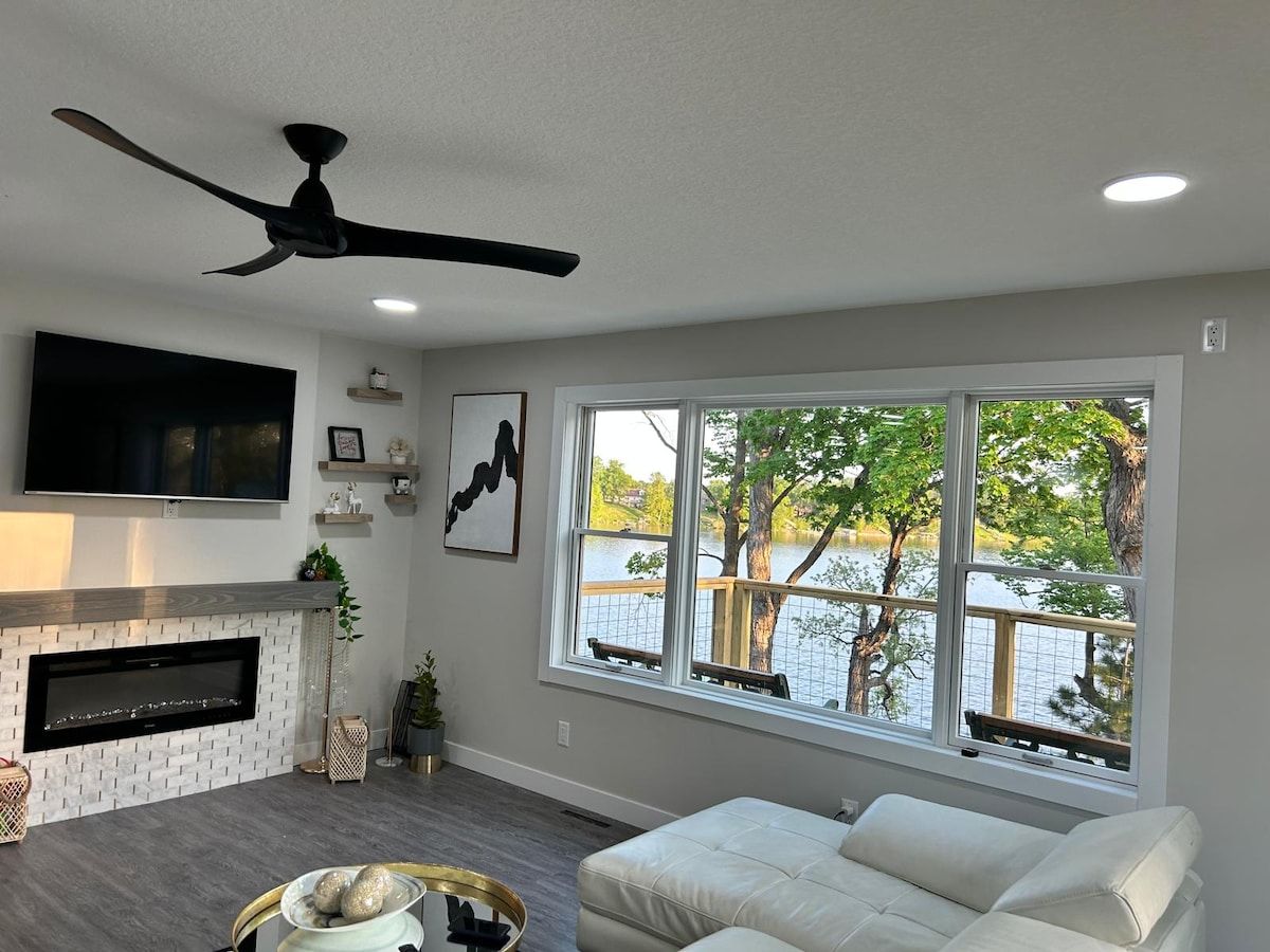 Ein Wohnzimmer mit Kamin, Deckenventilator, großem Fenster mit Seeblick und einem weißen Sofa.