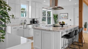 Una cocina moderna con armarios blancos, una isla grande con taburetes de bar, una campana extractora de acero inoxidable y un ventanal que proporciona luz natural.