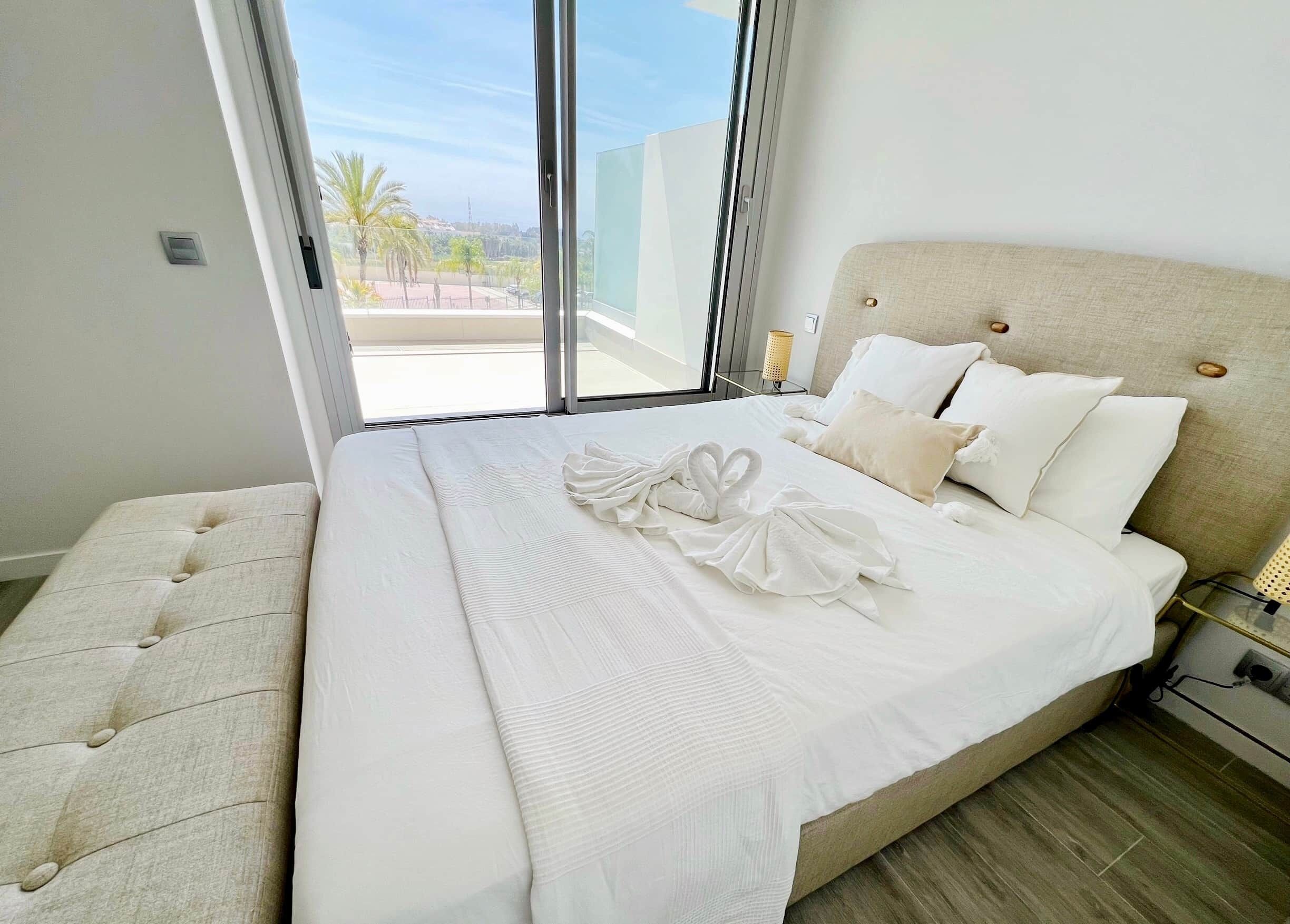 Una cama bien tendida con sábanas blancas y un juego de toallas, un cabecero y vistas a palmeras a través de un ventanal grande.