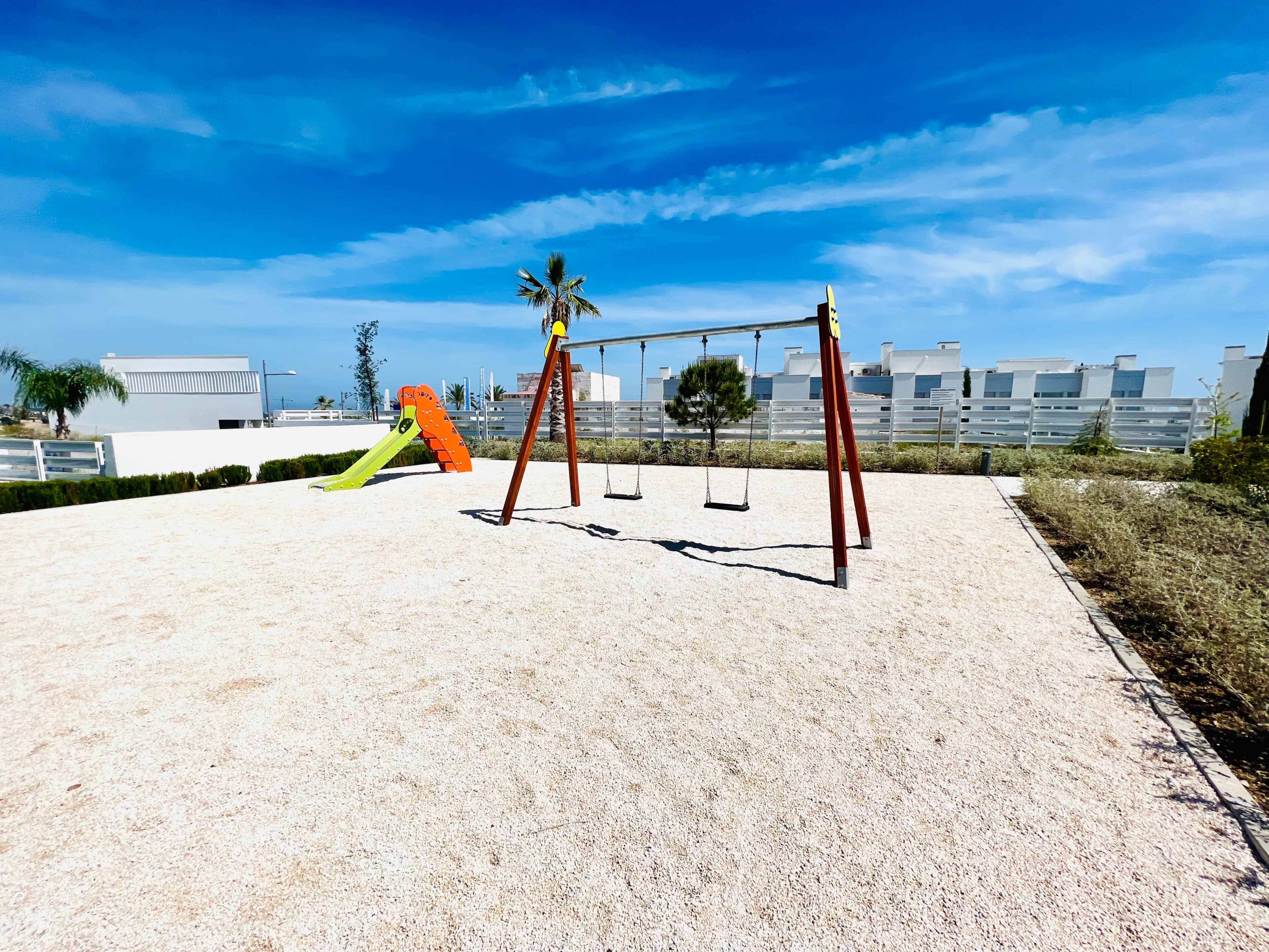 A playground with swings and a slide, surrounded by a sandy area and buildings in the background.