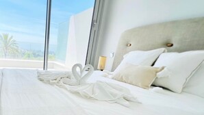 Et hotelværelse med en stor seng, hvide sengetøj, en sengegavl og udsigt til palmer gennem vinduet.
