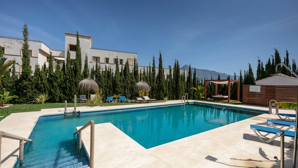 A swimming pool with lounge chairs and a wooden structure, surrounded by trees and a building with a modern design. - RDR301 - PH in Golden Banús with Sea and Mountain views (Marbella)