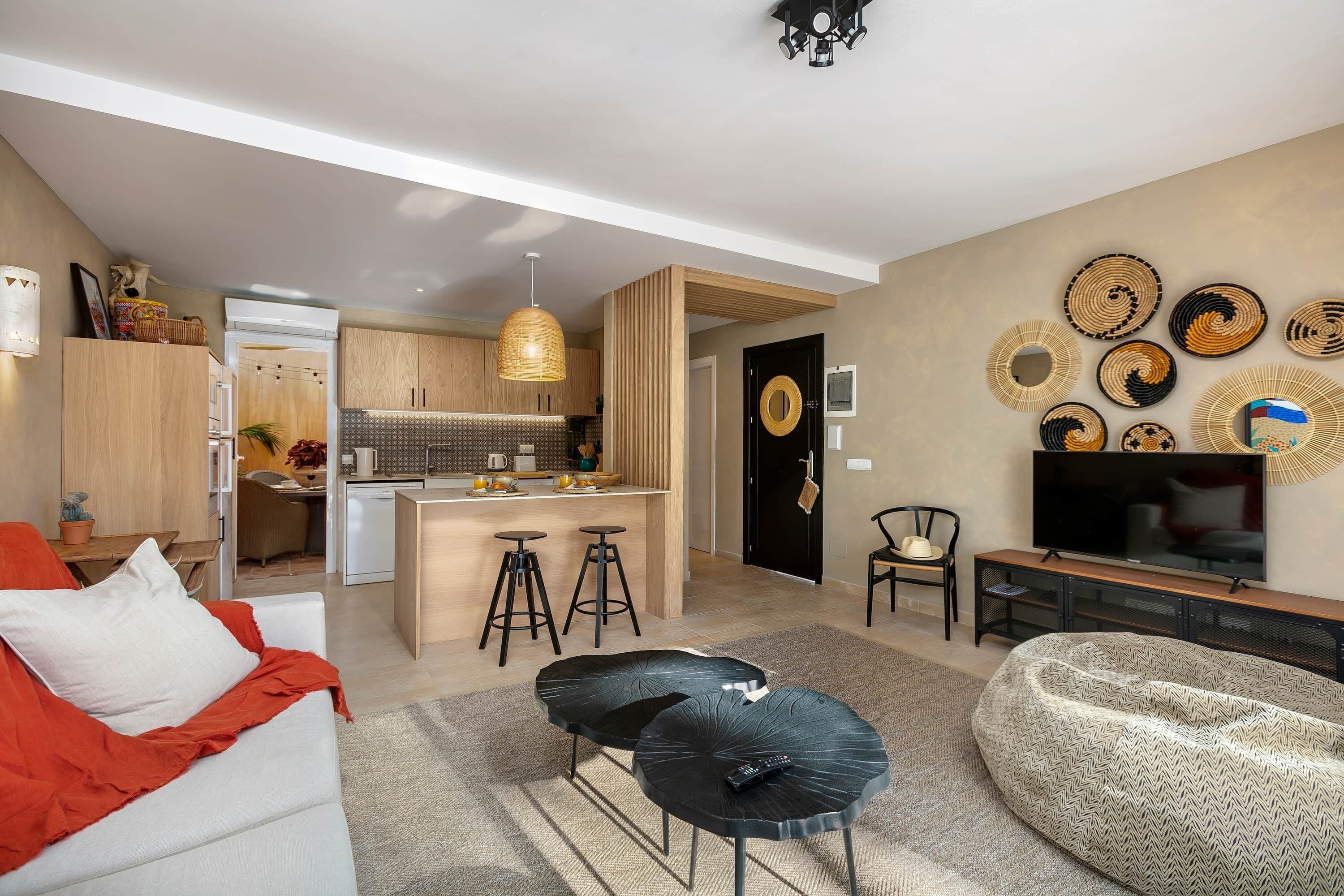 A modern living room with a sofa, ottoman, and a TV. There is a kitchen area with stools and a dining table.