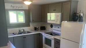 Cucina con pensili grigi, un frigorifero bianco, un forno a microonde e un fornello. Si nota una finestra sopra il lavello e un lampadario sopra il piano cottura.