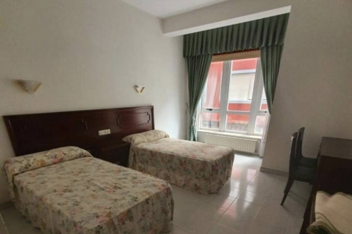 A room with two single beds, a wooden headboard, a small table, and a window with curtains.