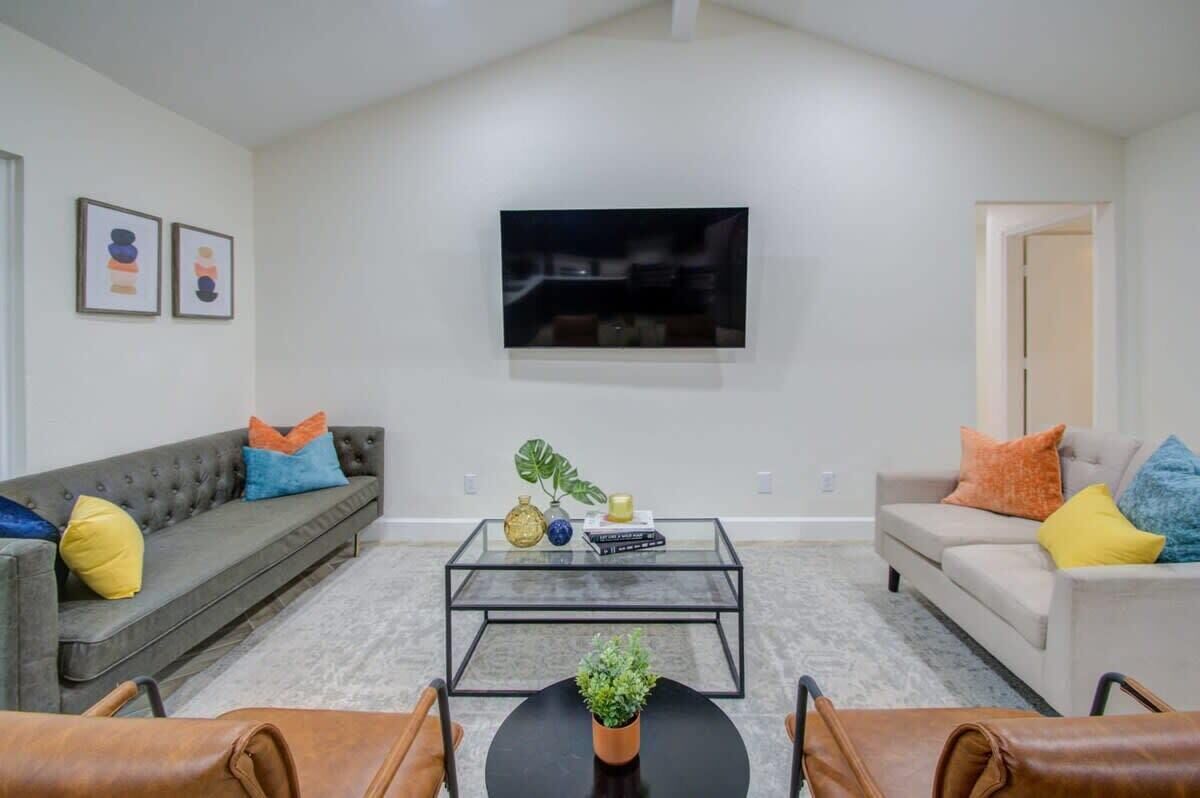 Een woonkamer met een grijze bank, een beige bank, een glazen salontafel en een aan de muur bevestigde televisie.