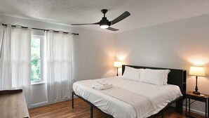 A neatly made bed with white linens, a ceiling fan, two bedside lamps, and a window with sheer curtains.