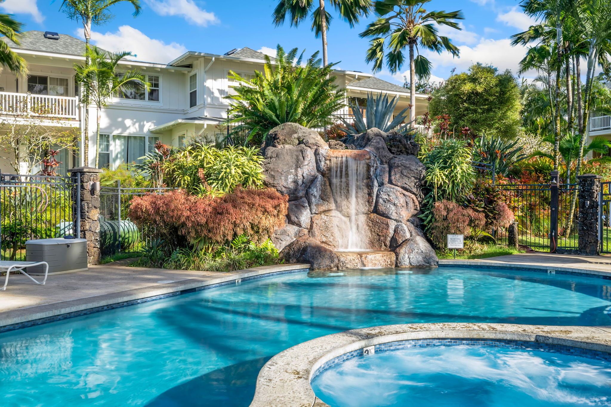 A pool with a waterfall feature, surrounded by tropical plants and palm trees, with a building in the background.