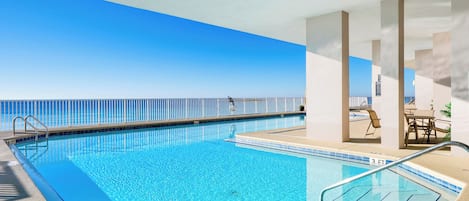 A clear blue pool with a view of the ocean, a covered seating area with tables and chairs, and a lifeguard chair.