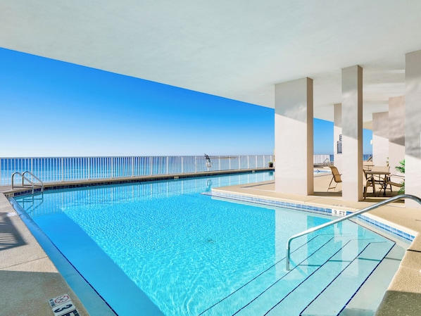 A clear blue pool with a view of the ocean, a covered seating area with tables and chairs, and a lifeguard chair. - Windemere #1202 | Gulf Views + Pool & Hot Tub! (Perdido Key)