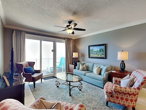 A hotel room with a balcony, a ceiling fan, a sofa, a coffee table, and a TV. - Windemere #1202 | Gulf Views + Pool & Hot Tub! (Perdido Key)