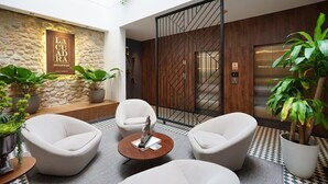 A modern lobby with a stone wall, a wooden panel wall, and a checkered floor. There are three white armchairs, a round table, and potted plants.
