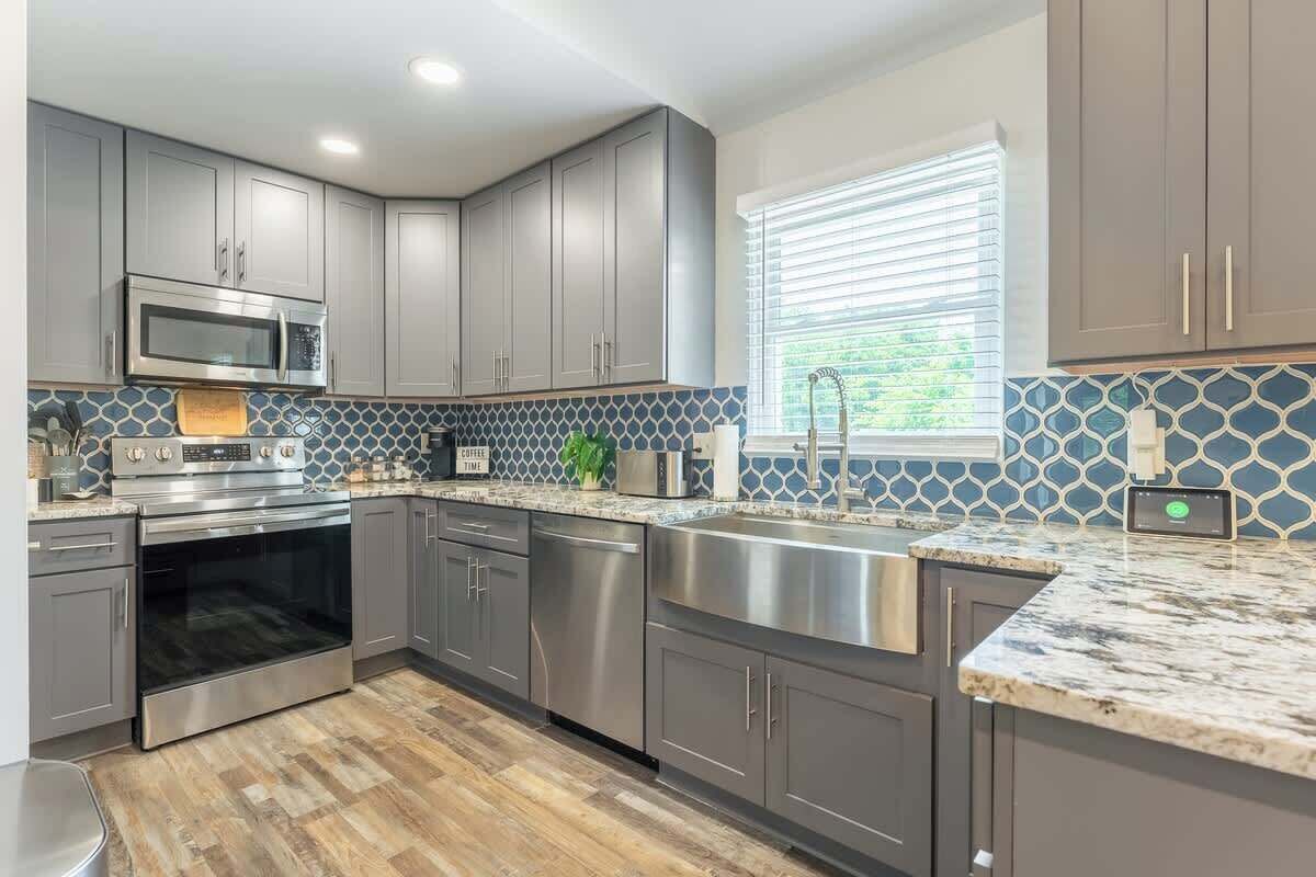 Una cocina moderna con armarios grises, electrodomésticos de acero inoxidable, salpicadero de azulejos y una ventana con persianas.