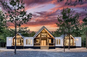 Situated in a natural setting, this cabin features a central entrance and two smaller wings, with large windows offering expansive views of the surrounding landscape.