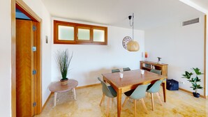 Un coin repas avec une table et des chaises en bois, une petite table d’appoint avec une plante, une bibliothèque et une horloge décorative au mur.