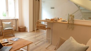 A cozy room with a wooden table, wicker chairs, a sofa, and a small kitchen area with a counter and stools.
