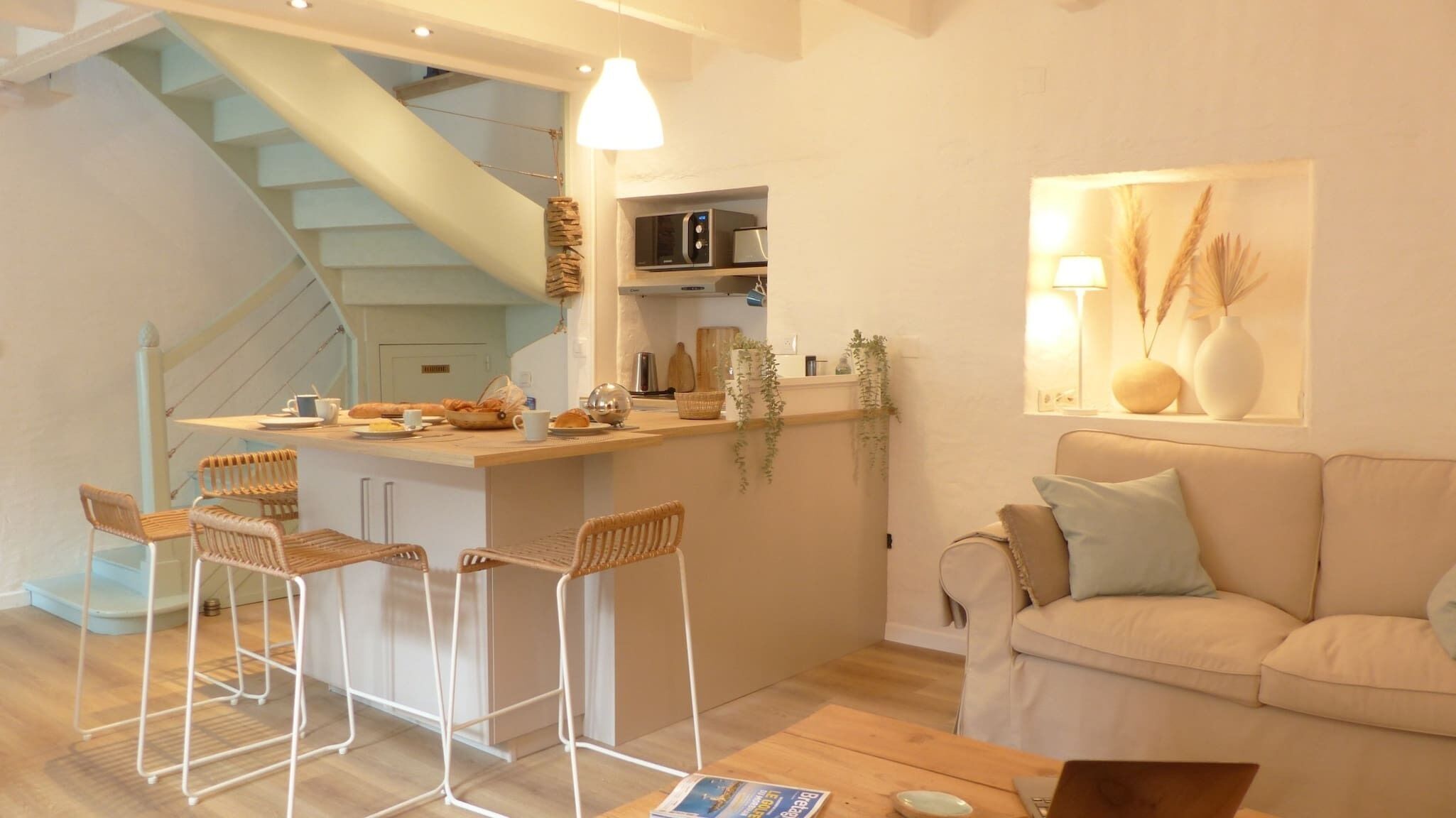 A modern kitchen with a central island, bar stools, a staircase, and a living area with a sofa and a lamp.