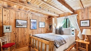 Un dormitorio tipo cabaña con cama, una silla, una mesita de noche y una ventana con cortinas.