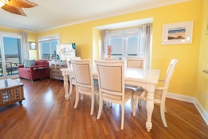 A dining area with a white table and chairs, a red sofa, and a view of the ocean through large windows.