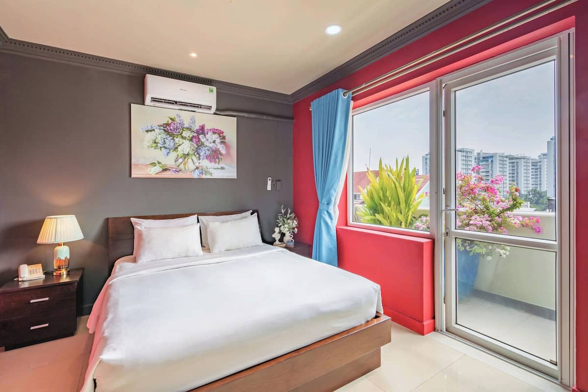 A hotel room with a bed, bedside table, lamp, air conditioning unit, and a large window with a view of buildings and plants.