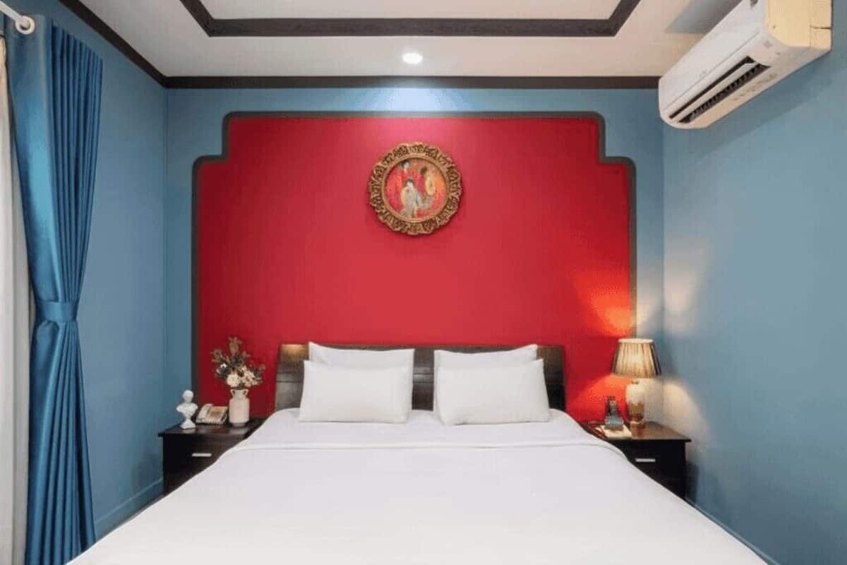 A hotel room with a red accent wall, a bed with white linens, a nightstand with a lamp, and an air conditioning unit.