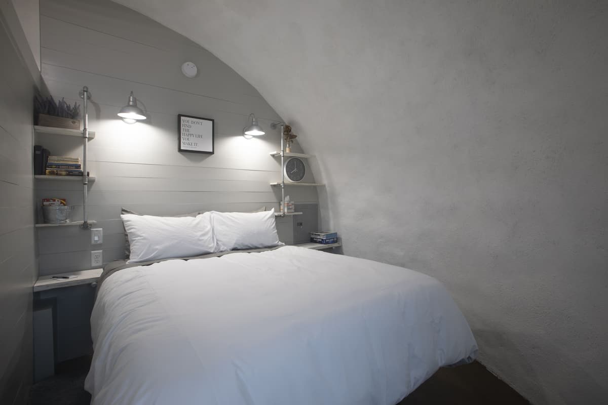 A bedroom with a bed, bedside tables, a wall clock, shelves with books and decorative items, and a ceiling light fixture.