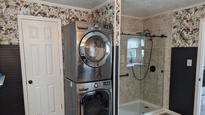 A compact laundry room with a stacked washer and dryer, a glass shower enclosure, and a door with a patterned wallpaper. - Little Blue Farmhouse (Jonesville)