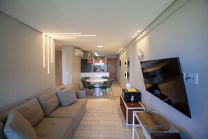 A modern living room with a sofa, a dining table with chairs, a TV mounted on the wall, and a kitchen in the background. - Flat de Luxo em Muro Alto (Porto de Galinhas Ipojuca)