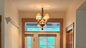 Ein Flur mit Doppeltüren, einer Chandelüster, Holzdielen und einem Fenster mit Blick auf Grünflächen.