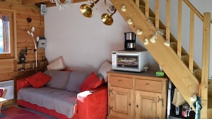 A cozy room with a wooden staircase, a sofa with red cushions, a small cabinet, and a microwave on top of it. - Appartement Plein Sud (Les Deux Alpes)